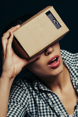 Color shot of a young woman looking through a cardboard, a device with which one can experience virtual reality on a mobile phone.の写真素材