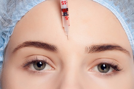 Botox injection. Attractive young woman in medical headwear and sketches on face keeping eyes closed while doctors hand making an injection in faceの写真素材