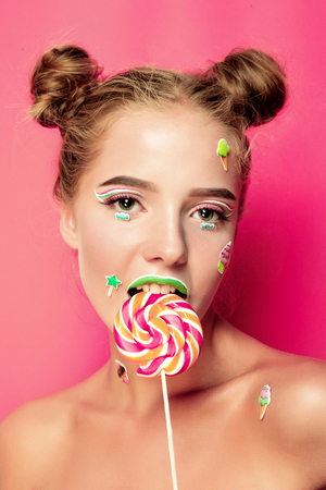 Beautiful young woman holds in hands candy smiling broadly. Stylish girl with bright makeup and candy in her hands, isolated on pink backgroundの写真素材