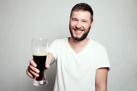 Drunk tattooed bearded man in a white T-shirt isolated on a grey background. A guy with a glass. smiling man, happyの写真素材