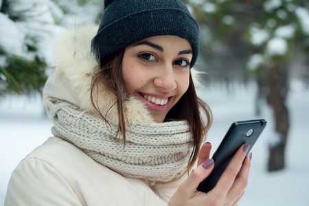 Pretty brunette girl doing a selfie with her smart phone.Beautiful young woman. Outdoor winter portrait. beautiful smiling girl talking on cell phone in snowy winter parkの写真素材