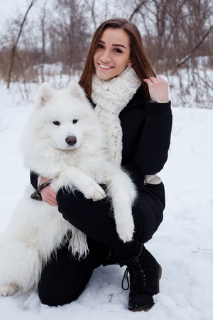 winter, christmas, animal, people, beauty concept- happy young woman owner petting embracing white Samoyed dog on snowの写真素材