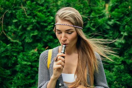 technologies, beauty and lifestyle concept -Happy vaping young white blonde girl. smoking fruit flavored e-liquid or e-juice with vaporizer device or e-cig.Modern gadget for smokersの写真素材
