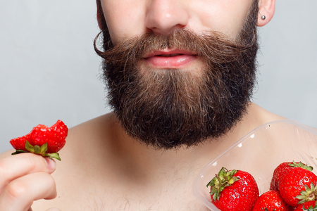 people, food, beauty, lifestyle, valentines day concept -close-up portrait young man holding a strawberry and smiling. Portrait of handsome bearded and mustache. on a gray backgroundの写真素材