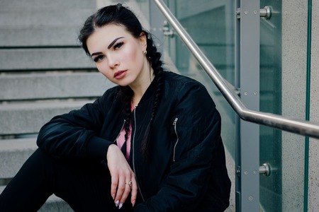 People, beauty, fashion, lifestyle concept -Young sexy woman with braids dressed in a black pants and a black bomber jacket. Fashion girl sits on the steps of a business building. Outdoors lifestyle portraits, hiphop styleの写真素材