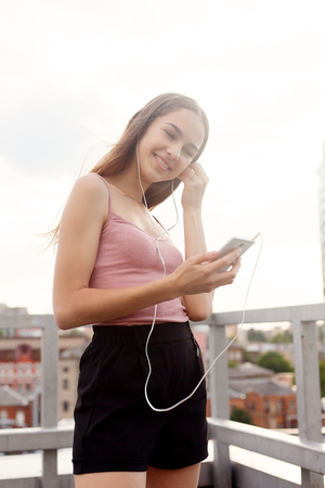 technologies, emotions, people, music, beauty, fashion and lifestyle concept - Young woman with headphones dangling on her mobile phone as she walks in an urban street, view from a heightの写真素材