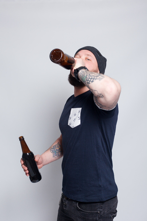 people, 4K and lifestyle concept - Brutal bearded male with tattooed arm drinks a beer from a bottle. handsome man in hat.Brutal bearded boy with tattooの写真素材