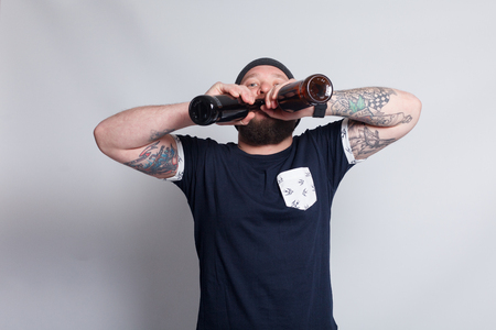 people, 4K and lifestyle concept - Brutal bearded male with tattooed arm drinks a beer from a bottle. handsome man in hat.Brutal bearded boy with tattooの写真素材