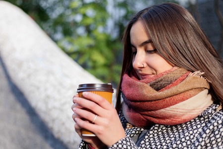 Portrait of beautiful brunette girl with takeaway drink on the street. scarf and autumn coat in the city. Amazing view of business lady with a cup of coffee walking down the city street.の写真素材