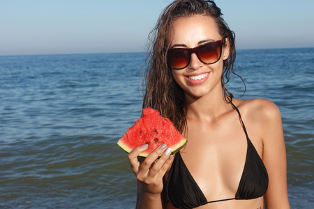 Holiday, resort, tourism concept - Summer vacation - young girl eating fresh watermelon on sandy beach. young beautiful woman eats watermelon on the beach at hot summer day.の写真素材