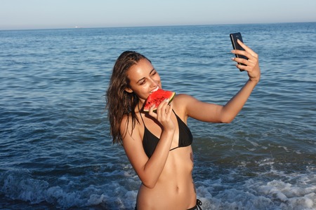 Holiday, resort, tourism concept - Summer vacation - young girl eating fresh watermelon on sandy beach. young beautiful woman eats watermelon on the beach at hot summer day.の写真素材