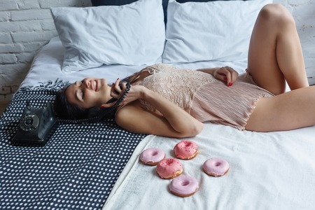 people, sweets and fast food concept - happy young woman or teen girl eating donut, kneeling in bed....
