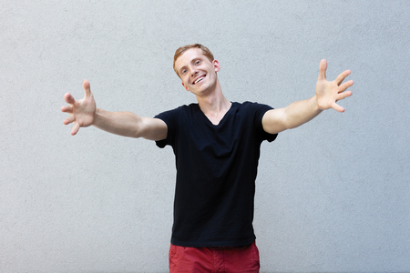 fashion, style, emotions and people concept - Close up portrait of a redhead of a beautiful manly guy with freckles on a gray background black T-shirt. pulls his arms to embrace with a smileの写真素材