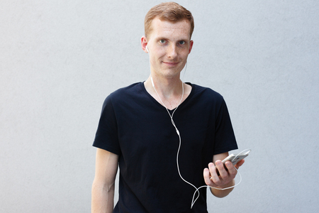 fashion, emotions and people concept - Close up portrait of a redhead beautiful manly guy. frecklessongs on smart phone with earphones, dances, gestures happily, being inspired by favourite musicの写真素材