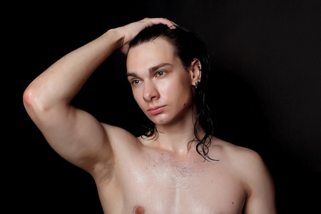 people, skincare and beauty concept - Wet young man with long black hair on a black background. Portrait male with shaved chest . Men's skin care. Ripped muscular handsome man on black backgroundの写真素材