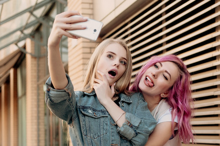 Friends taking selfie with a smart phone and making faces and fun. Closeup of girls making funny faces and smiling for selfie. Two happy girlfriends taking photo with their smartphone in the cityの写真素材