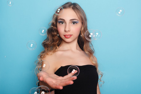 Beautiful young woman with soap bubbles over cyan background. Portrait of young beautiful girl in...