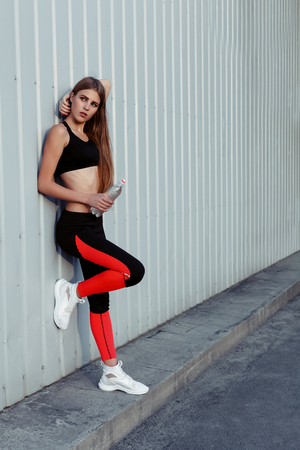 Female athlete drinking water while standing by a grey wall. Slim woman in sportswear taking a break after workout.の写真素材