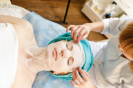 Collagen face mask . Facial skin treatment. Woman receiving cosmetic procedure in beauty salon. Bottle with moisturizing cream on background.の写真素材