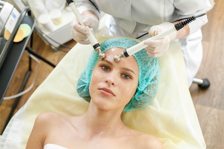 people, beauty, spa, cosmetology and technology concept - Cosmetologist makes the procedure microcurrent therapy of the facial skin of a beautiful, young woman in a beauty salonの写真素材