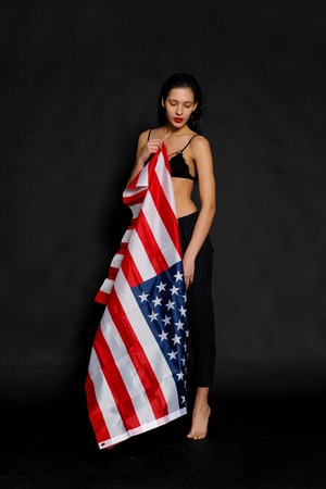 Portrait of proud female athlete wrapped in American Flag against black background. Muscular young woman looking confidently at camera.の写真素材