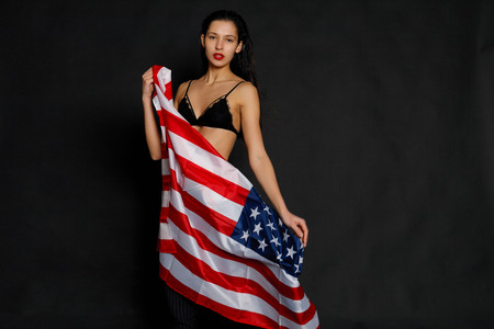 Portrait of proud female athlete wrapped in American Flag against black background. Muscular young woman looking confidently at camera.の写真素材