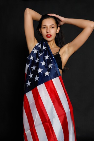 Portrait of proud female athlete wrapped in American Flag against black background. Muscular young woman looking confidently at camera.の写真素材