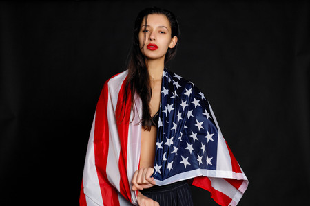 Portrait of proud female athlete wrapped in American Flag against black background. Muscular young woman looking confidently at camera.の写真素材