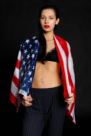 Portrait of proud female athlete wrapped in American Flag against black background. Muscular young woman looking confidently at camera.の写真素材