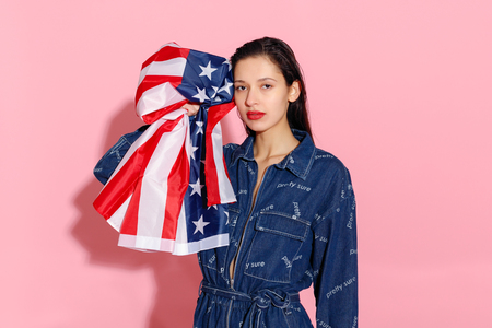 Portrait of proud female athlete wrapped in American Flag against pink background. Muscular young woman looking confidently at camera. female protesting for equality and women empowermentの写真素材