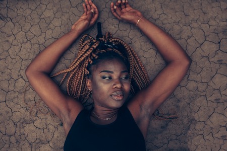 Beautiful African female wearing black dress lying on dry cracked ground in the desert. water shortage, dryness, diseaseの写真素材