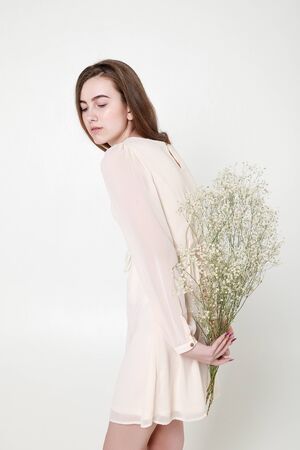 Curly romantic look, beautiful eyes. White wildflowers in hands. Girl white light dress and curly hair, portrait of woman with flowers on white background, purity and innocenceの写真素材