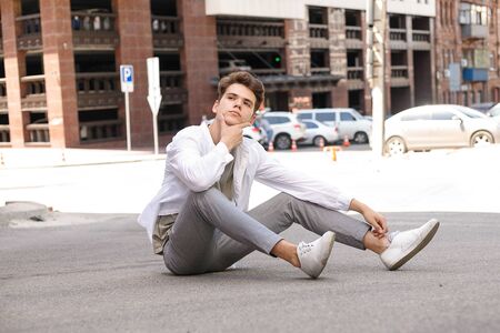guy model with a stylish haircut posing outdoors in a white shirt and gray trousers. trendy hairstyle rests near a modern business center. Attractive guy in the street on a summer day.の写真素材