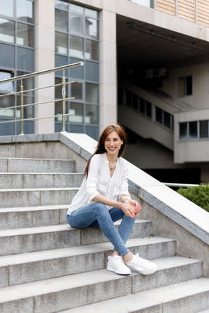 Attractive young woman in the city. Business lady standing near business center. Tired business woman with white shirt in blue jeans sitting relax on the street near city centre glass buildingの写真素材