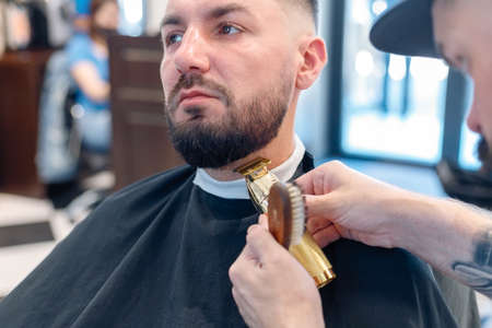 Hairstyling process. Close-up of a barber drying hair of a young bearded man. Young man in barber shop sits in an armchair. Barber cuts his hair with a trimerの写真素材