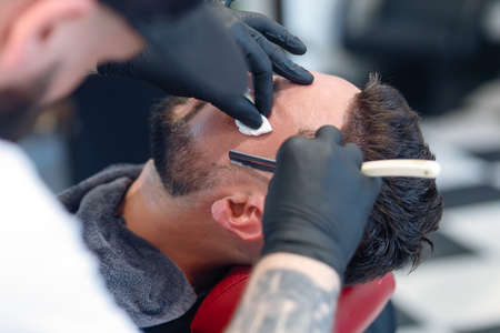 Professional barber shaves customer beard with straight razor. Beard cut with old-fashioned blade at...