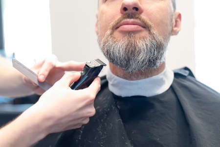 Hairstyling process. Close-up of a barber drying hair of a young bearded man. Young man in barber shop sits in an armchair. Barber cuts his hair with a trimerの写真素材