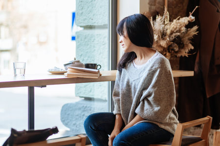 Young woman drinking coffee and reading book sitting indoor in urban cafe. Cafe city lifestyle. Casual portrait of teenager girl. Toned.の写真素材