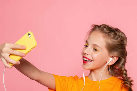 Cute little girl taking selfie. Photo of cute lovely small lady long telephone show thumb-up make selfie toothy beaming smile wear casual stylish red t-shirt isolated pink color backgroundの写真素材