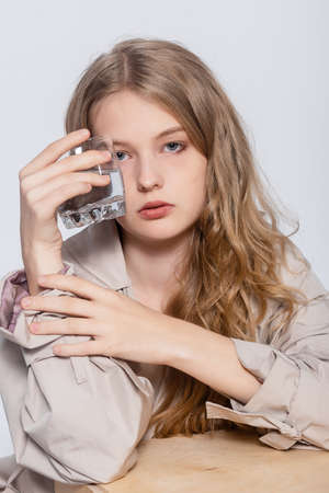 Smiling Young Woman with glass of Water, beautiful girl in beige raincoat posing in Studio. Smiling business woman drink water. White background. Isolated portrait.の写真素材