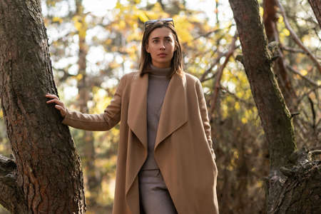 Stylish young lady in a long beige coat and black hat walking by the river in a city green parkの写真素材