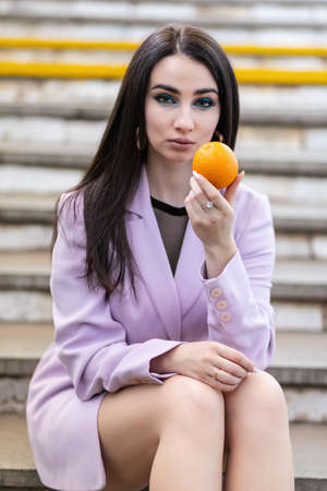 Fashion pretty woman model with fruit oranges sits on the steps in a purple jacket, black shoes.の写真素材
