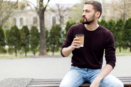 Half length portrait of bearded blogger in stylish sunglasses strolling on street with coffee to go and smartphone in hands. Cheerful hipster guy dressed in casual wear checking notification on phoneの写真素材