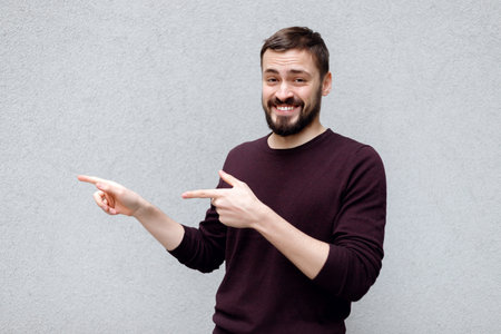 Handsome adult spouse with beard and mustache, smiling and expressing cheer while pointing left with two hands, standing against gray background. Artist offers to show his new work to close friendsの写真素材