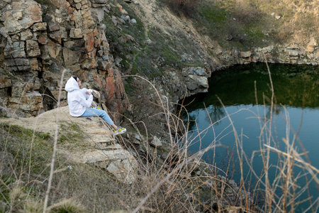 girl sits on the edge of a mountain in a white hoodie and looks at the lake, Mountain and coastal...