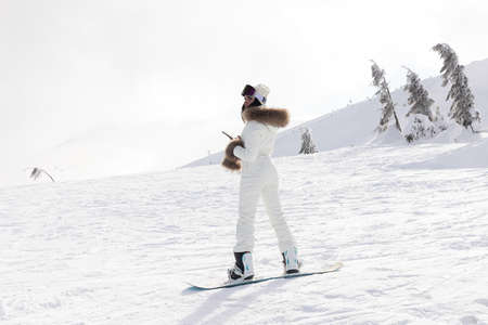 winter, leisure, sport and people concept - Girl snowboarder stands with snowboard on mountain's top on sunrise backdrop. girl holding a phone with her backの写真素材
