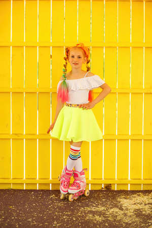 Little girl roller skating against yellow background, wearing shirt and leggins, touching her hair, looking directly at camera, cute female child having fun.の写真素材