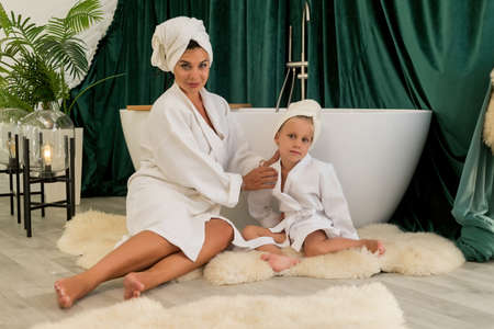 home spa. Mother and daughter make a clay facial mask. Beautiful young women with child doing beauty treatment together. Cute girl with mom in towel in the bathroom.の写真素材