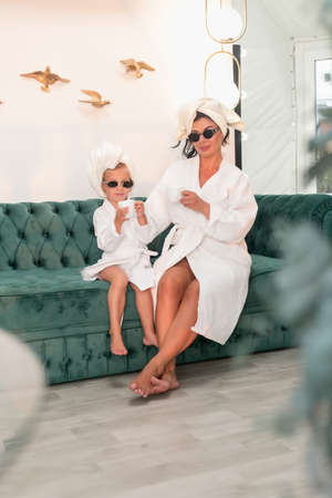 Mother with child doing beauty treatment together. Happy family mother and child daughter in towel and bathrobe are sitting on the sofa and drinking teaの写真素材