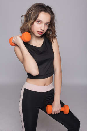 Happy young woman with dumbbell in hand making exercises outdoors in front of gray wall, looking smiling directly at camera, training arms, wearing white top and beige leggins.の写真素材
